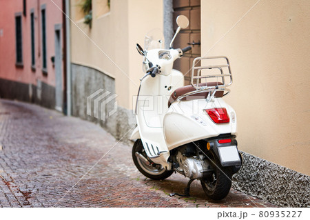 White retro scooter or moped parked on sidewalk of empty narrow street. Motorcycle or motorbike on italian street White retro scooter or moped parked on sidewalk of empty narrow street. Motorcycle or motorbike on italian street 80935227