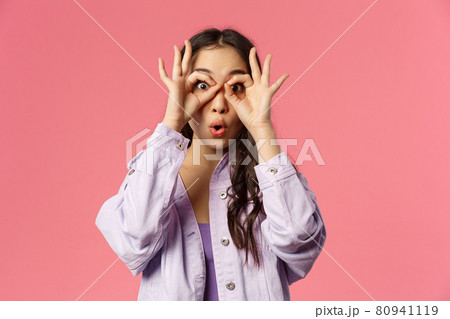 Close-up portrait of amused, speechless and impressed asian young girl seeing something curious and amazing, make glasses with fingers over eyes, fold lips say wow astonished, pink background Close-up portrait of amused, speechless and impressed asian young girl seeing something curious and amazing, make glasses with fingers over eyes, fold lips say wow astonished, pink background 80941119