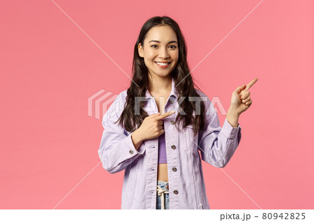 Portrait of cheerful asian girl, female student inviting people follow page, subscribe to page or visit link, pointing fingers right and smiling camera, standing happy over pink background 80942825