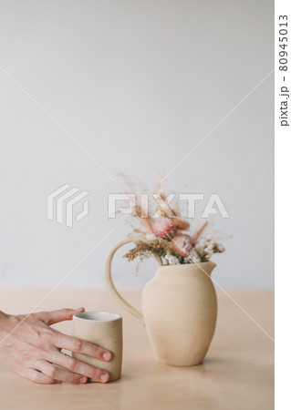 Minimal home decor with dry flowers in a ceramic vase and a cup in female hands. Minimal home decor with dry flowers in a ceramic vase and a cup in female hands. 80945013
