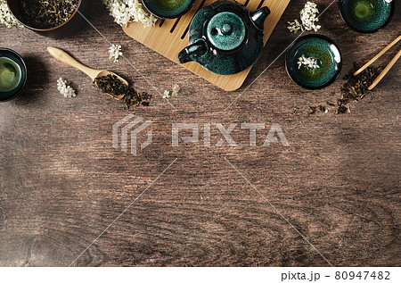 Dark turquoise glazed clay teapot and small cups on dark wooden background decorated with white flowers. Creative minimalist tea ceremony concept. Copy space, top view. 80947482