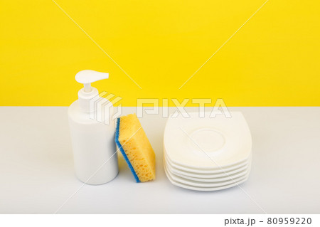 Dishwashing detergent in white bottle, pile of saucers and cleaning sponge against yellow background 80959220