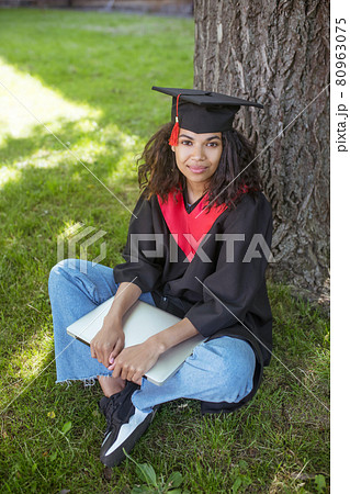 A girl in academic gown sitting under the tree with a laptop 80963075