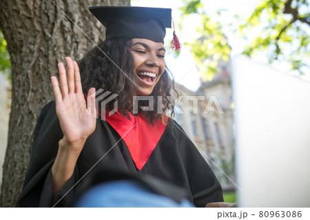 A girl in academic gown sitting under the tree with a laptop 80963086
