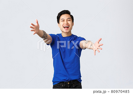Cheerful friendly-looking young male student, asian man in blue t-shirt with tattoos, reaching hands towards camera for hug, want cuddles or embraces, greeting dear friend, grey background 80970485