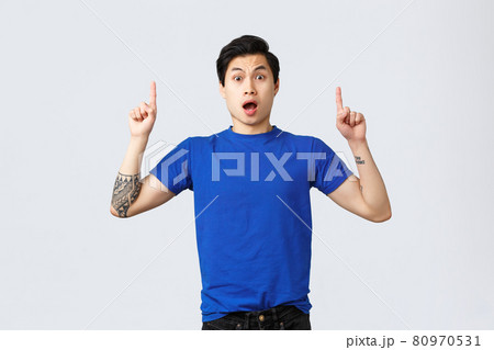 Different emotions, people lifestyle and advertising concept. Surprised and astonished asian male student, guy in blue t-shirt gasping amazed, pointing fingers up at banner, grey background Different emotions, people lifestyle and advertising concept. Surprised and astonished asian male student, guy in blue t-shirt gasping amazed, pointing fingers up at banner, grey background 80970531