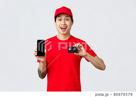 Delivery application, online orders and shipping concept. Quarantine wont stop from shopping. Cheerful asian courier in red cap and t-shirt, showing smartphone display and credit card 80970579