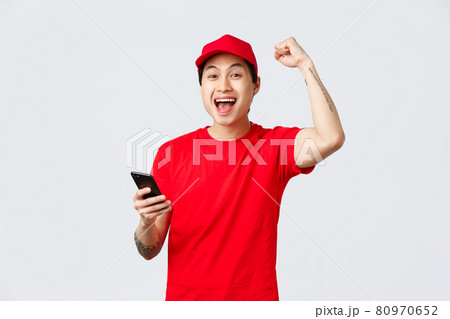 Happy, cheerful asian delivery guy in red uniform cap and t-shirt of carrier service, holding smartphone, reading good news, chanting with fist pump, yell yes celebrate victory or achivement 80970652