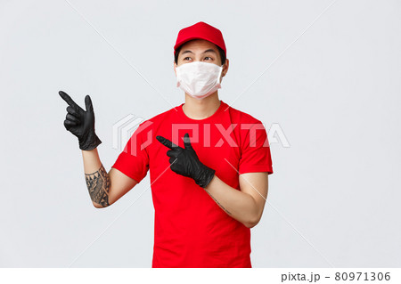 Enthusiastic smiling asian delivery man in medical mask and protective gloves, looking pointing upper left corner, showing courier service new promo or special offer, wearing red uniform Enthusiastic smiling asian delivery man in medical mask and protective gloves, looking pointing upper left corner, showing courier service new promo or special offer, wearing red uniform 80971306