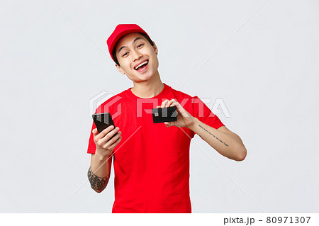 Friendly delivery guy in red uniform t-shirt and cap, showing smartphone screen and credit card, recommend application for tracking order online shopping. Courier advertise carrier service 80971307