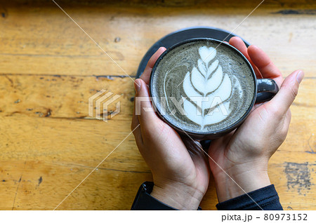Someone hands holding a cup of Goth Latte or Charcoal Latte on wooden table (Focus on coffee surface). The black colour is caused by adding activated charcoal to espresso and then adding milk. 80973152