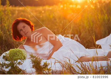 Charming woman in white bed in a field at sunset in summer looking at the camera 81003996