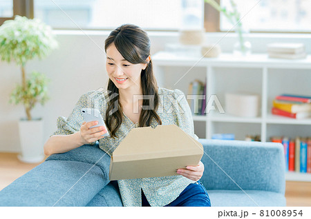 リビングのソファーに座りスマホを見ながら荷物を確認する若い主婦 81008954