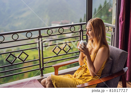 A young woman holding coffee cup while sittinhg on chair on balcony , looking at mountains and green nature. Woman in Sapa in the fog, Northwest Vietnam. Vietnam travel concept. UNESCO heritage A young woman holding coffee cup while sittinhg on chair on balcony , looking at mountains and green nature. Woman in Sapa in the fog, Northwest Vietnam. Vietnam travel concept. UNESCO heritage 81017727