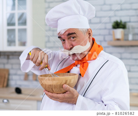 An old man with a bushy gray mustache in a chef's hat is cooking in the kitchen. An old man with a bushy gray mustache in a chef's hat is cooking in the kitchen. 81026582