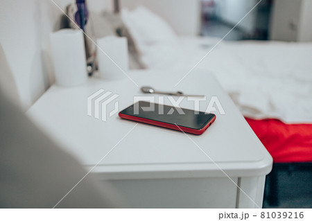Black smartphone in a red case on the bedside table. Women's watch on the table in the bedroom against the background of the bed. Close up 81039216