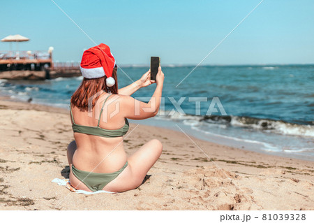 Young beautiful girl in a red santa claus hat and swimsuit makes a selfie on the seashore 81039328
