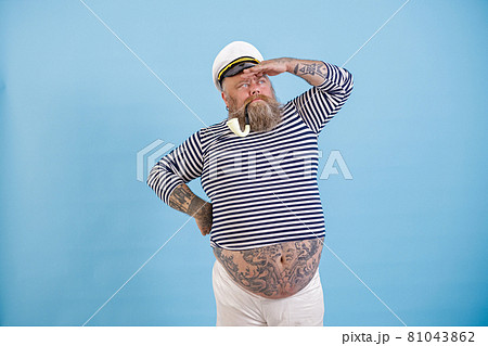 Plus size sailor with smoking pipe and hat looks into distance on light blue background Plus size sailor with smoking pipe and hat looks into distance on light blue background 81043862