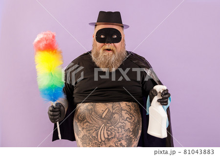 Emotional plump man in hero suit holds brush and spray bottle on purple background 81043883