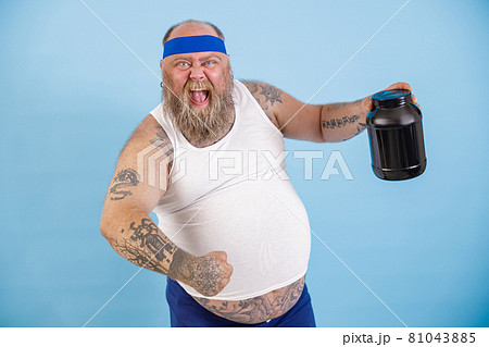 Joyful bearded man with overweight holds black bottle of protein supplement on light blue background 81043885