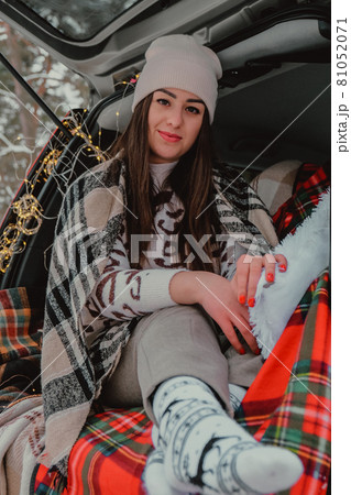 Brunette woman wrapped in blanket in trunk car drinking coffee tea from thermos. Travel in winter. Car decorated with festive Christmas lights. Outdoor picnic Brunette woman wrapped in blanket in trunk car drinking coffee tea from thermos. Travel in winter. Car decorated with festive Christmas lights. Outdoor picnic 81052071