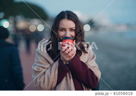 Young beautiful woman in coat and scarf with coffee cup outdoor. 81056019