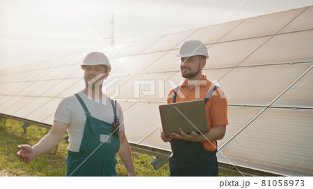 Male engineers standing at solar farm and discussing efficient plan of construction. Two men in protective helmet. The engineer holds his hand in the air, a place for a virtual screensaver. Male engineers standing at solar farm and discussing efficient plan of construction. Two men in protective helmet. The engineer holds his hand in the air, a place for a virtual screensaver. 81058973