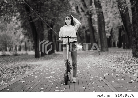 Caucasian female wearing protective face mask riding scooter in city park during covid pandemic. Caucasian female wearing protective face mask riding scooter in city park during covid pandemic. 81059676