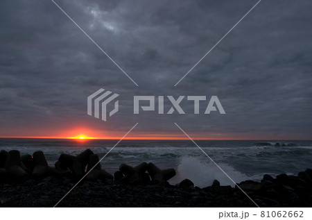 低く立ち込める雲の水平線近くの切れ間からオレンジ色の太陽が輝く、荒れた灰色の海に光明が印象的で綺麗だ 81062662