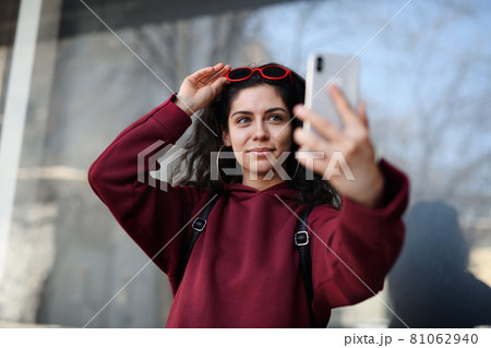 Portrait of young woman with smartphone standing outdoors on street in city, taking selfie. 81062940