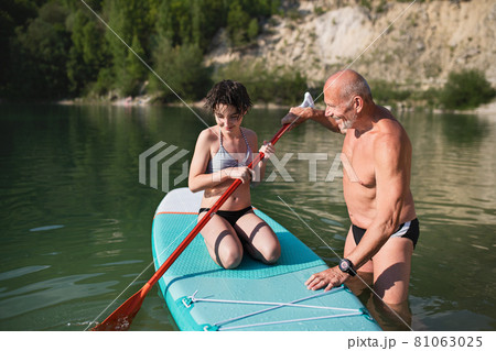 Happy preteen girl with grandfather on summer holiday by lake, paddleboarding. 81063025