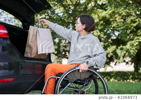 Woman sitting at her wheelchair and opening car trunk and putting shopping bags inside Woman sitting at her wheelchair and opening car trunk and putting shopping bags inside 81068973