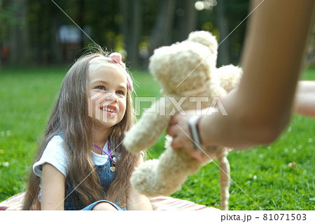 Happy child girl playing with her teddy bear toy sitting on green grass in summer park. Happy child girl playing with her teddy bear toy sitting on green grass in summer park. 81071503