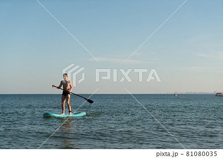 A 12 year old boy learns to stand on a SUP board in the sea near the shore. 81080035