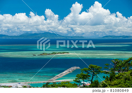 Stunning top view of a tropical island, pier extends over the blue and green gradient sea, blue sky, white clouds over Ishigaki island on horizon, green vegetation. Iriomote Island. Stunning top view of a tropical island, pier extends over the blue and green gradient sea, blue sky, white clouds over Ishigaki island on horizon, green vegetation. Iriomote Island. 81080849