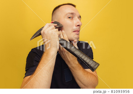 One young brutal man, firefighter shaving with an axe over yellow studio background. 81093956