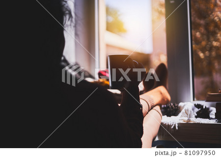 A cozy autumnal day. A woman hanging her legs on the window sill. Cold autumn sunny day. Drink coffee near the window A cozy autumnal day. A woman hanging her legs on the window sill. Cold autumn sunny day. Drink coffee near the window 81097950