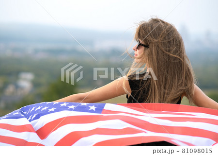 Young pretty american woman with long hair holding waving on wind USA flag on her sholders standing outdoors enjoying warm summer day. Young pretty american woman with long hair holding waving on wind USA flag on her sholders standing outdoors enjoying warm summer day. 81098015
