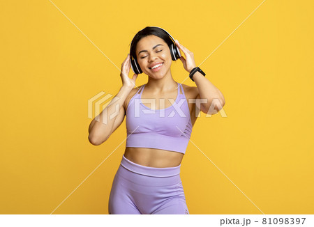 Favourite song. Excited black sportive lady in wireless headphones listening to music and smiling, yellow background Favourite song. Excited black sportive lady in wireless headphones listening to music and smiling, yellow background 81098397