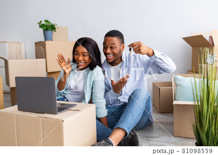 Excited black couple chatting online with friends and showing key of their house to camera, sitting among carton boxes Excited black couple chatting online with friends and showing key of their house to camera, sitting among carton boxes 81098559