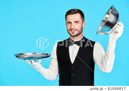 Elegant young waiter holding cloche over empty tray ready to serve on blue background Elegant young waiter holding cloche over empty tray ready to serve on blue background 81099817