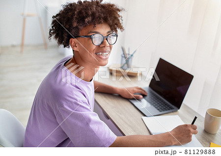 Happy black adolescent sitting at desk with laptop, learning remotely, taking notes in copybook at home Happy black adolescent sitting at desk with laptop, learning remotely, taking notes in copybook at home 81129888