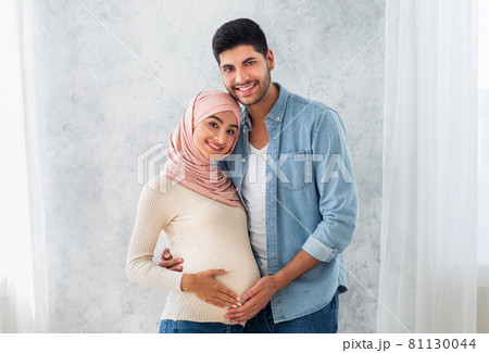 Portrait of young pregnant arab spouses, embracing and smiling at camera, loving muslim man cuddling his wife's belly Portrait of young pregnant arab spouses, embracing and smiling at camera, loving muslim man cuddling his wife's belly 81130044