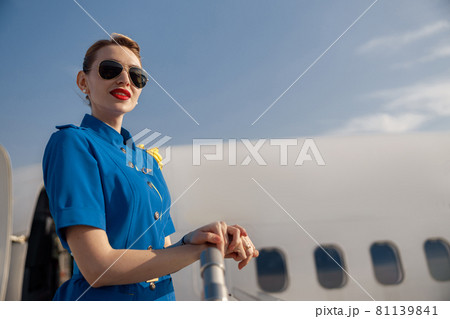 Portrait of pretty air stewardess in blue uniform and sunglasses looking away, standing on airstair on a daytime 81139841
