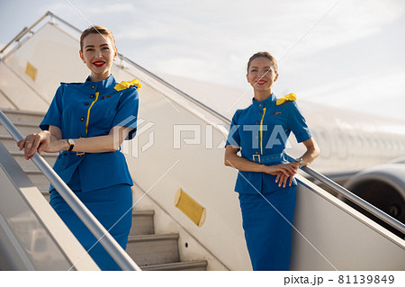 Two beautiful air stewardesses in blue uniform smiling at camera, standing on airstair and welcoming passengers Two beautiful air stewardesses in blue uniform smiling at camera, standing on airstair and welcoming passengers 81139849