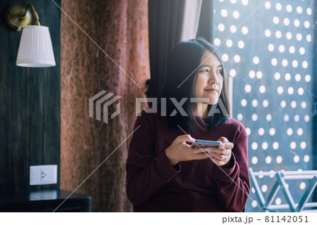 Young smiling asian woman sitting on the bed at home and looking on window with smartphone,Positive thinking concept Young smiling asian woman sitting on the bed at home and looking on window with smartphone,Positive thinking concept 81142051