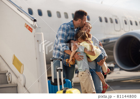 Family of four kissing each other while going on a trip, standing in front of big airplane outdoors 81144800