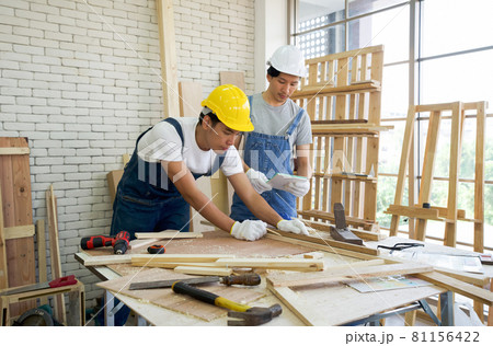 An Asian carpenter is drawing a line on a piece of wood, preparing to cut into sections. The other man told the size he wanted by looking at his note. Morning work atmosphere in the workshop room. 81156422