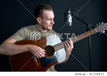 Young caucasian man sings while playing an acoustic guitar in front of black soundproofing walls. Musicians producing music in professional recording studio. 81156651