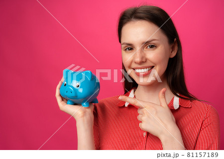 Happy smiling young woman holding blue piggy bank against pink background 81157189